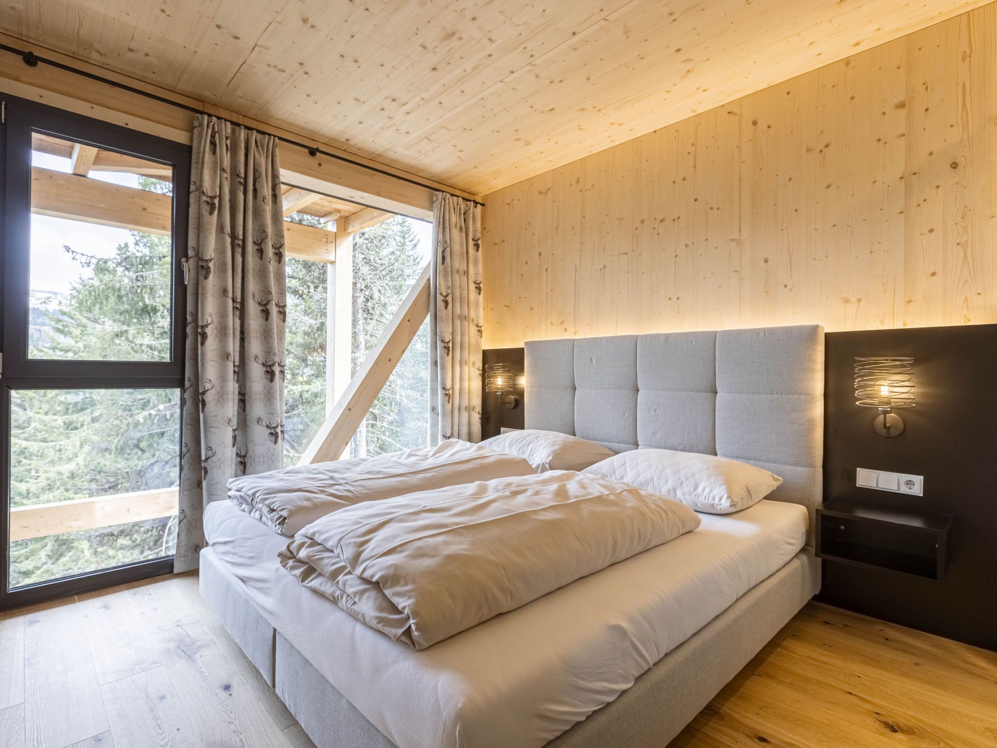 A bedroom with a large bed, wooden walls, and a view of trees through a window.