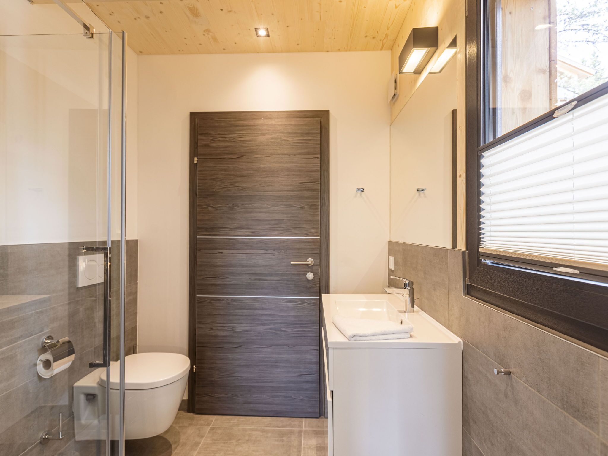 A modern bathroom with a glass shower enclosure, a dark wooden door, and a white sink with a towel.