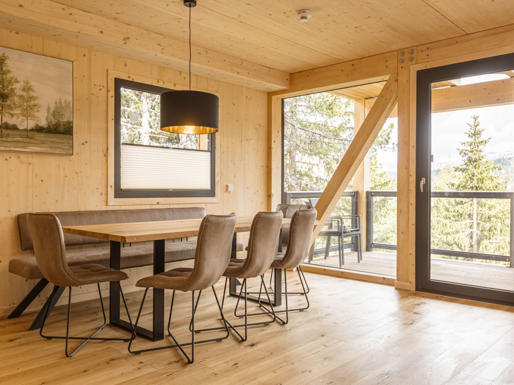 A wooden interior with a dining table and chairs, a pendant light, and a large window with a view of trees.