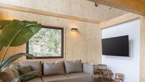 A modern living room with a sectional sofa, a flat-screen TV mounted on the wall, and a large window providing a view of trees.