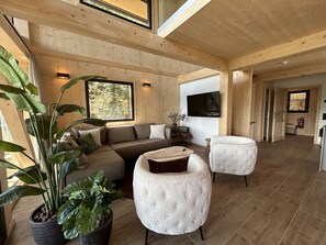 Een ruime woonkamer met een grote bank, een rond poef en een aan de muur bevestigde televisie.