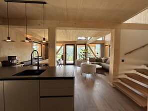 Un intérieur moderne comprenant une cuisine, un coin repas et un espace de vie, avec des poutres en bois, de grandes fenêtres et un escalier.