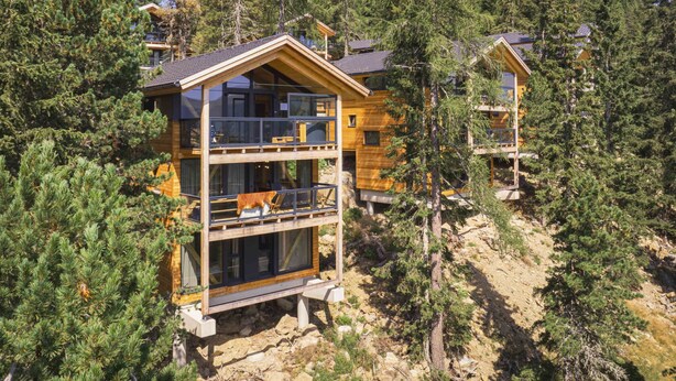 A multi-story wooden cabin with a balcony, surrounded by dense forest.