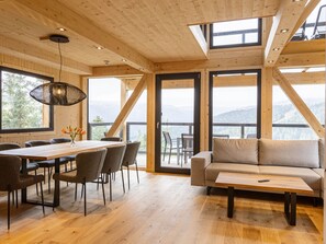 Ein geräumiges Zimmer mit Holzboden, einem Esstisch mit Stühlen, einer Couch und einem großen Fenster mit Blick ins Freie.