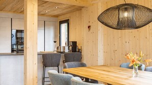 A modern kitchen with a wooden ceiling, a dining area with a wooden table and grey chairs, and a hanging light fixture.