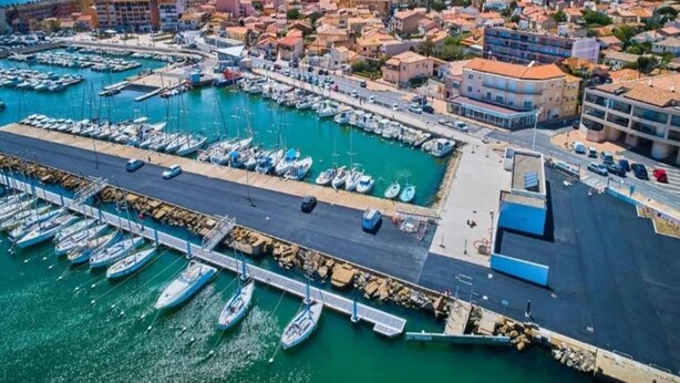 Vue aérienne d’une ville côtière avec un port de plaisance, des bâtiments et un ciel dégagé.