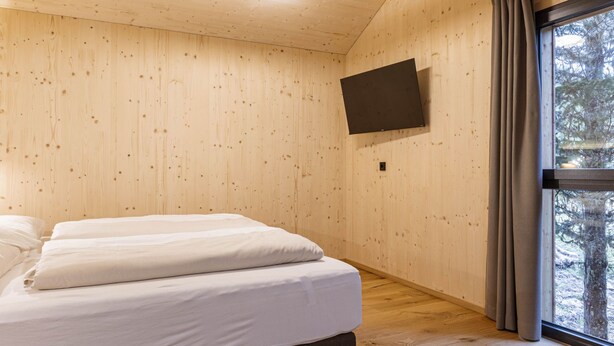 Een kamer met houten wanden, een bed met witte beddengoed, een wandtelevisie en een raam met gordijnen.