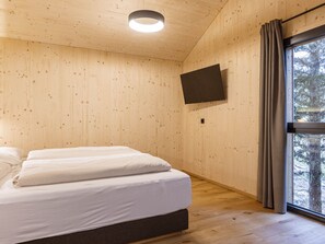 Quarto com paredes de madeira, cama com roupa de cama branca, TV de tela plana fixada na parede e janela com cortinas.