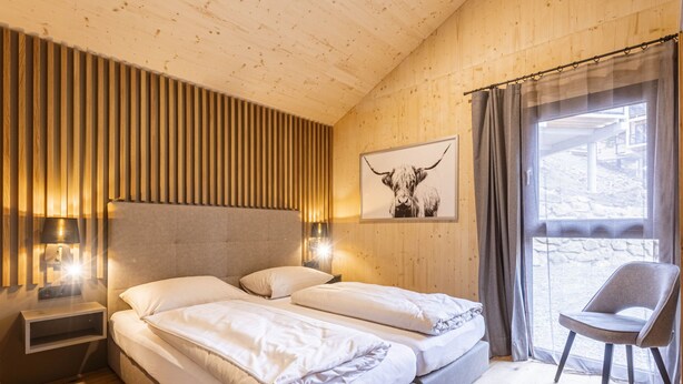 A bedroom with a wooden slat wall, a bed with white linens, a mounted picture of a bull, a chair, and a window with curtains. - 02_NTH019_Sup Chalet mit Sauna und Hot Tub 2 Etage by Interhome (Turracher Höhe)