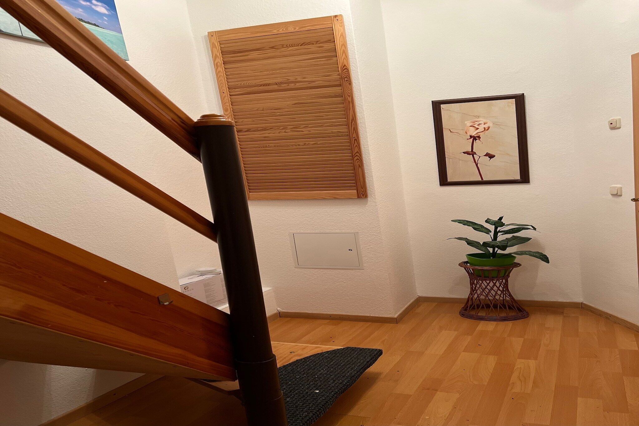 Eine Holztreppe mit Handlauf, ein Holzboden, ein an der Wand befestigtes Fenster, ein gerahmtes Rosenbild und ein Blumentopf.