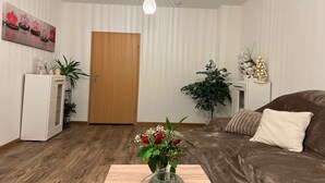 A living room with a brown sofa, a wooden door, a white cabinet, a green vase with flowers, and a ceiling light. - Airy City Apartment in Zwickau (Zwickau)