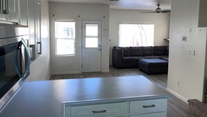 A modern kitchen with stainless steel appliances, a light-colored countertop, and a built-in oven. There is a gray sectional sofa in the living area, a door with a window, and a ceiling fan. - Charming 3-bedroom townhome with AC, WiFi in serene Ewa Beach (Ewa Beach)