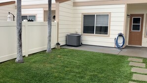 A residential backyard with a well-maintained lawn, a patio area, a window with blinds, and a door. - Charming 3-bedroom townhome with AC, WiFi in serene Ewa Beach (Ewa Beach)