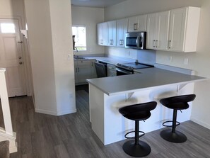 A modern kitchen with white cabinets, a stainless steel microwave, and a large island with two bar stools. - Charming 3-bedroom townhome with AC, WiFi in serene Ewa Beach (Ewa Beach)