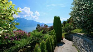 A stone pathway leading through a lush garden with various plants and a view of mountains in the background. - Magnificent villa with indoor pool and prime lake view location (Brione sopra Minusio)