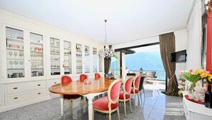 A dining area with a round table, red chairs, and a white cabinet with glass doors. - Magnificent villa with indoor pool and prime lake view location (Brione sopra Minusio)