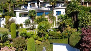 A large, modern house with a white facade, multiple balconies, and lush gardens. - Magnificent villa with indoor pool and prime lake view location (Brione sopra Minusio)