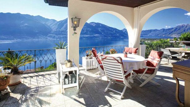 A terrace with a table and chairs, a potted plant, and a view of mountains and a lake. - Magnificent villa with indoor pool and prime lake view location (Brione sopra Minusio)
