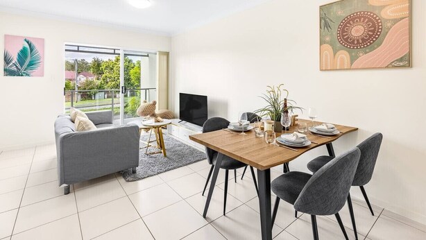 A modern living room with a grey sofa, a wooden dining table set for four, and a large window offering a view of greenery. - Walk to everything! Modern 2Beds Apt w/ EasyAccess (Springwood)