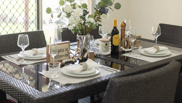 A dining table set with plates, glasses, a bottle, and a vase with flowers. - 3Beds Vibrant Home Steps to Mall & Transport (Sunnybank)