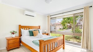 A bedroom with a wooden bed, a nightstand, a potted plant, and a large window with a view of greenery and buildings. - 3Beds Vibrant Home Steps to Mall & Transport (Sunnybank)