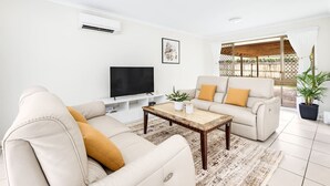 A living room with a sofa, a coffee table, a TV, and a window with a view of a wooden deck. - 3Beds Vibrant Home Steps to Mall & Transport (Sunnybank)