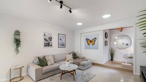 Ein modernes Wohnzimmer mit einem beigen Sofa, einem runden Couchtisch und einem grauen Teppich. An den Wänden befinden sich gerahmte Bilder, eine hängende Pflanze und ein runder Spiegel.