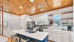 Uma cozinha moderna com armários brancos, uma ilha central com banquetas, eletrodomésticos de aço inoxidável e teto de madeira.