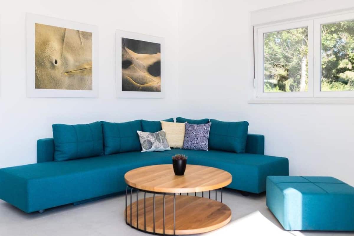 Ein blaugrünes Sofa mit einem runden Couchtisch in einem modernen Raum, an der Wand hängen zwei gerahmte Kunstwerke und ein Fenster mit Blick auf Bäume.
