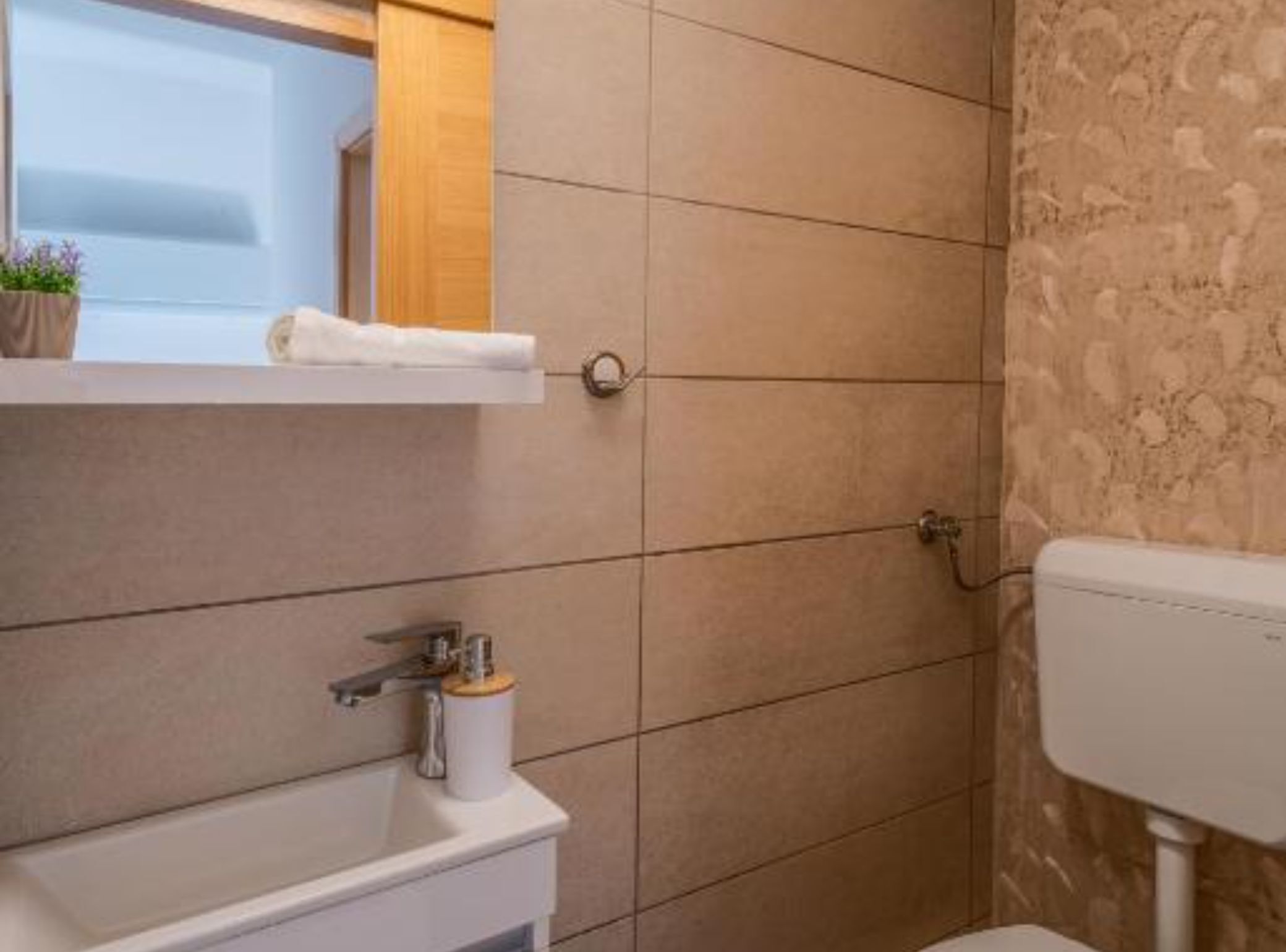 A bathroom with a white sink, a mirror, and a toilet. There is a shelf above the sink with a plant and a towel.
