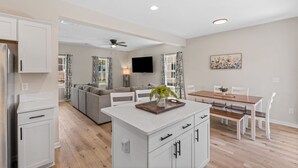 A modern kitchen with white cabinets, a dining area with a wooden table and chairs, a gray sofa, and a ceiling fan. - New House! Close to HPU & HPMKT (High Point)