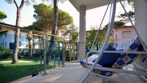 Un balcone con una sedia a dondolo, ringhiera in vetro e vista su un giardino e degli edifici.