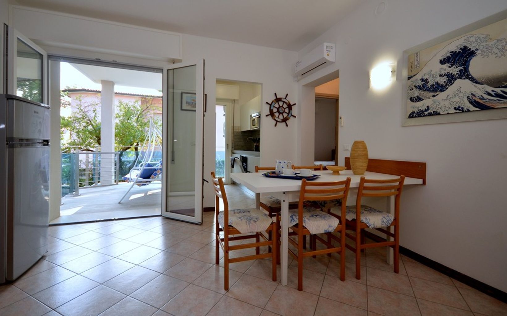 Zona pranzo con tavolo apparecchiato per quattro, sedie in legno, decorazione a forma di timone da nave e un'ampia finestra vista giardino.
