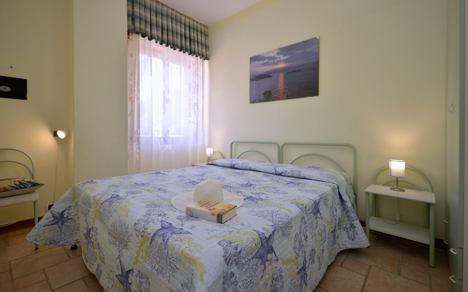 Un dormitorio con cama, una mesita de noche, una silla, una ventana con cortinas y un cuadro colgado en la pared.