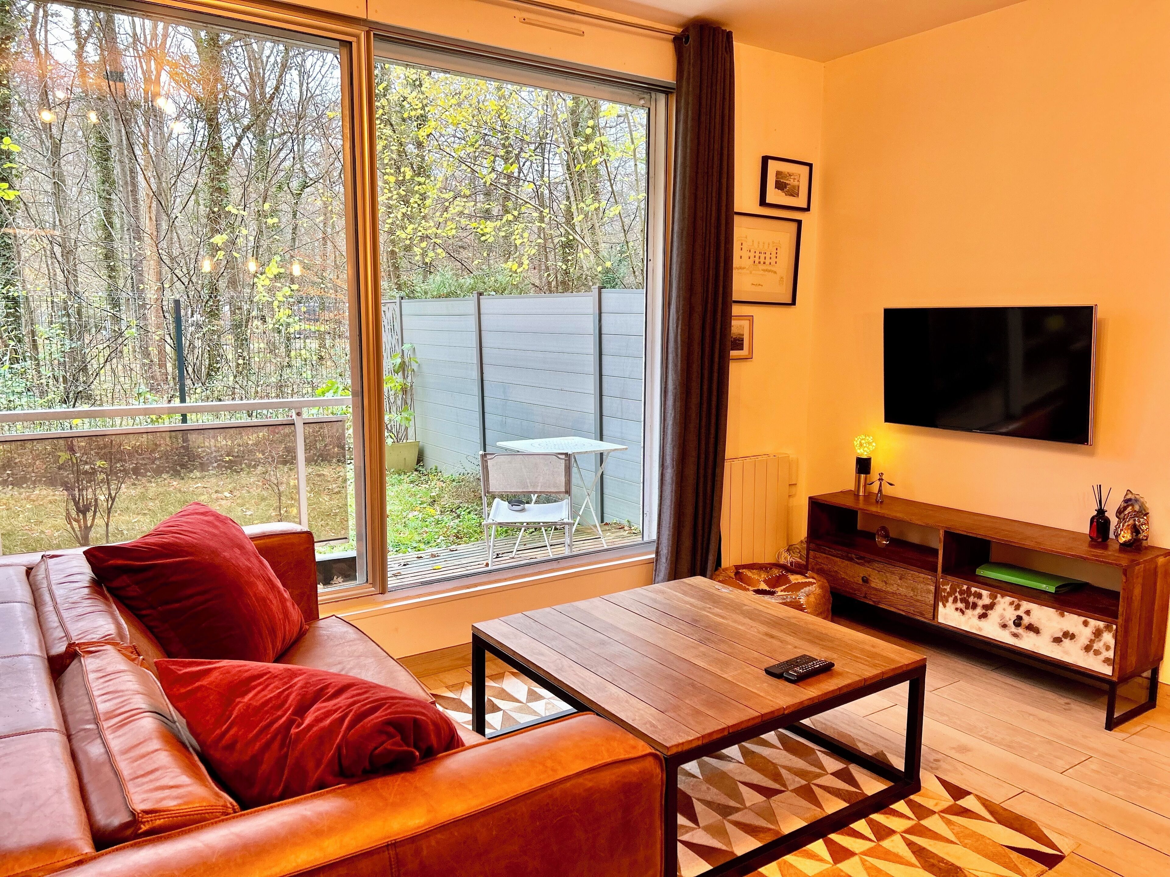 A living room with a leather sofa, a wooden coffee table, a TV, and a large window with a view of trees and a fence.