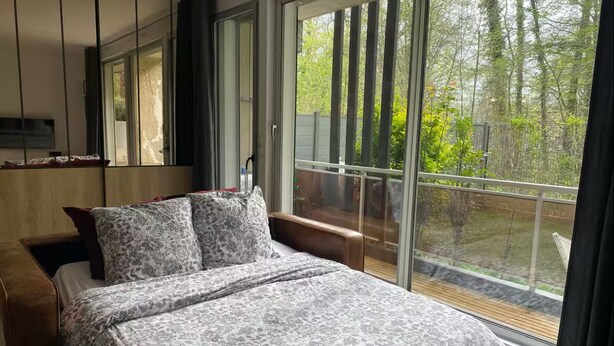 Un dormitorio con una cama grande, cabecera de madera, un espejo y vistas a un jardín a través de ventanas amplias.