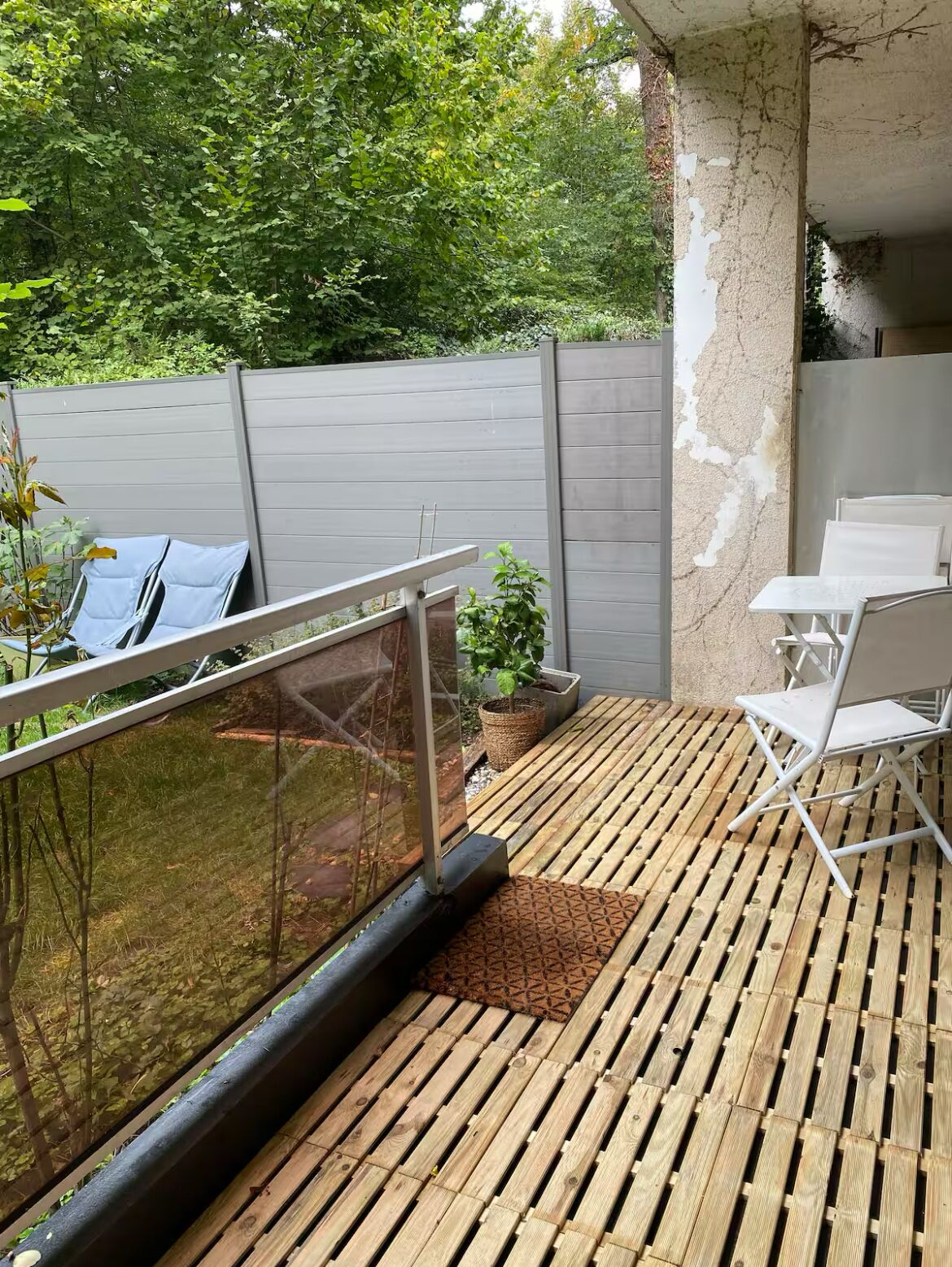 Una terraza de madera con barandilla de vidrio, una planta en maceta y un juego de mesa y sillas blancos.