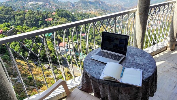 Un balcón con una laptop, un cuaderno y una silla, con vista a un paisaje pintoresco.
