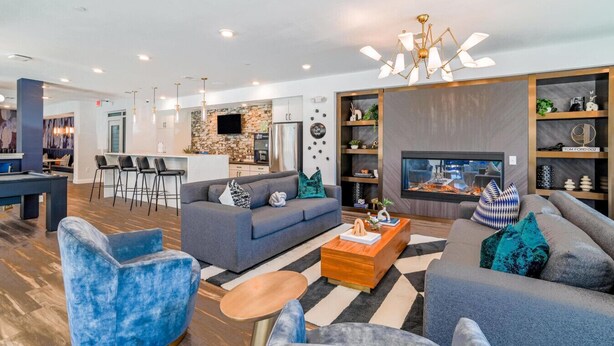 Una sala moderna con un sofá gris, un sillón azul y una mesa de centro. Se observa un área de cocina con salpicadero de piedra y un televisor de pantalla plana.