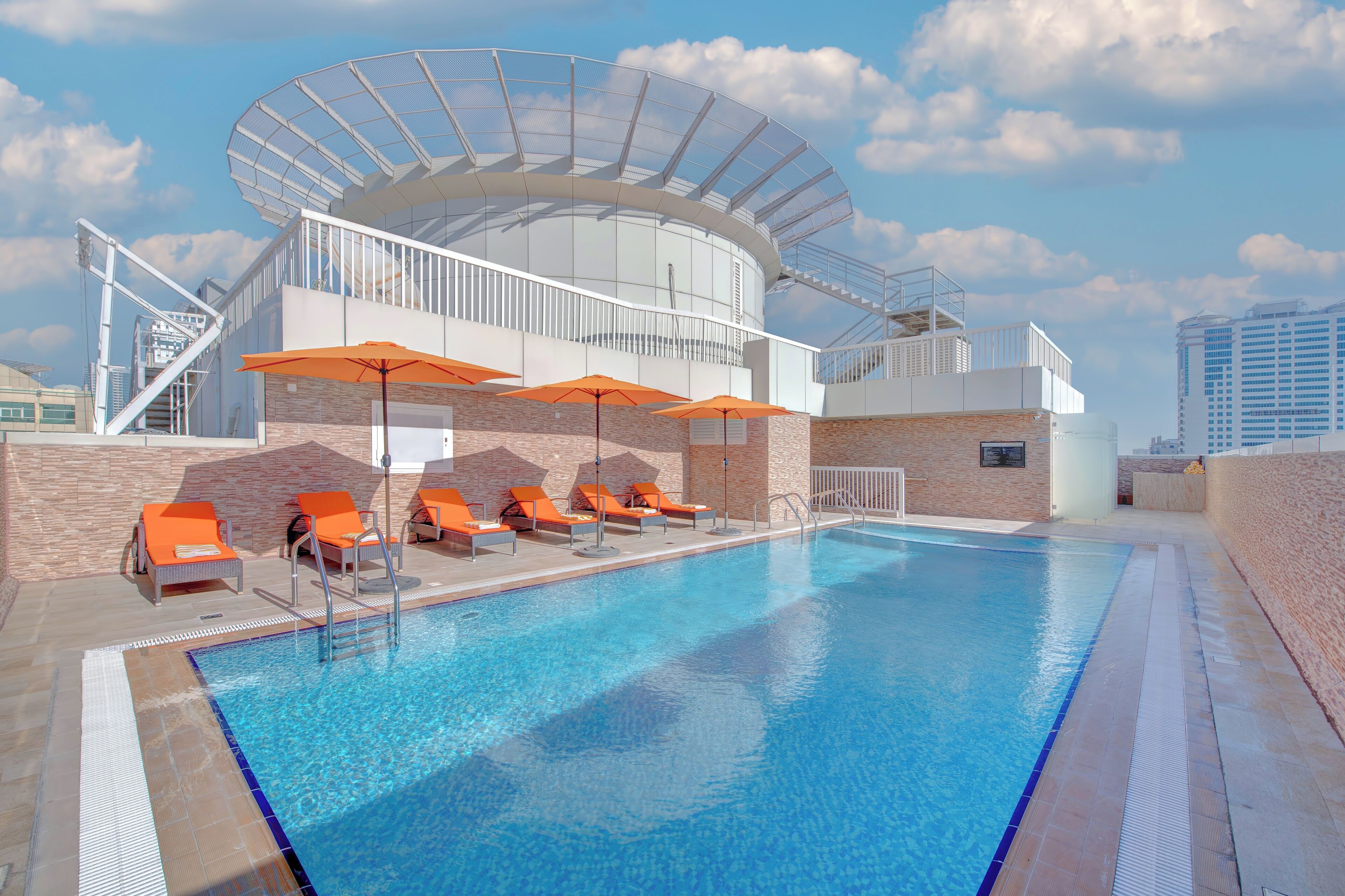 Una piscina con sillones de descanso y sombrillas, rodeada por un edificio moderno con techo de cristal y paredes de ladrillo.