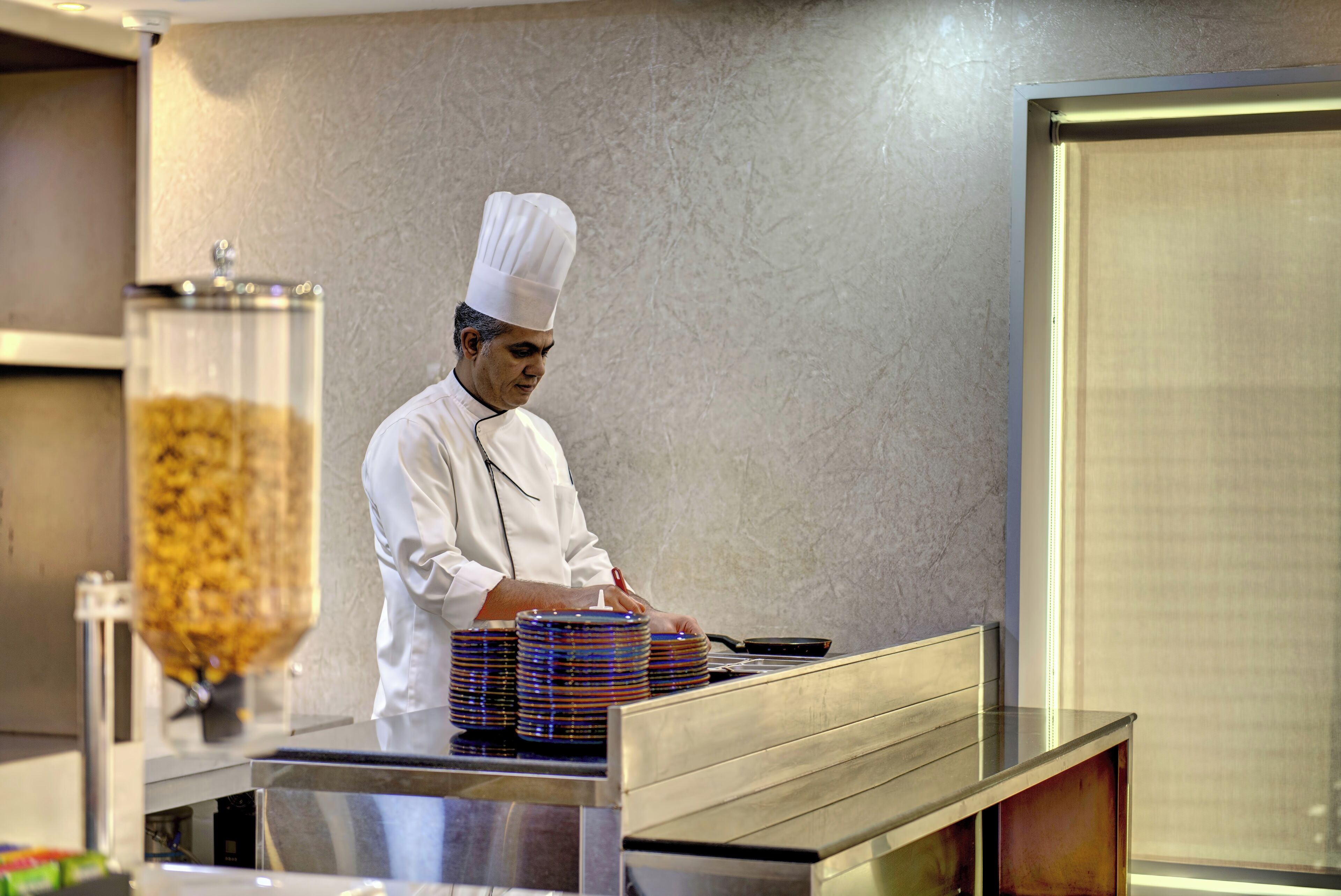A chef in a white uniform preparing food in a kitchen with a large window and a food dispenser.