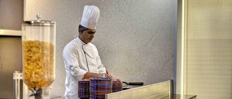 A chef in a white uniform preparing food in a kitchen with a large window and a food dispenser.