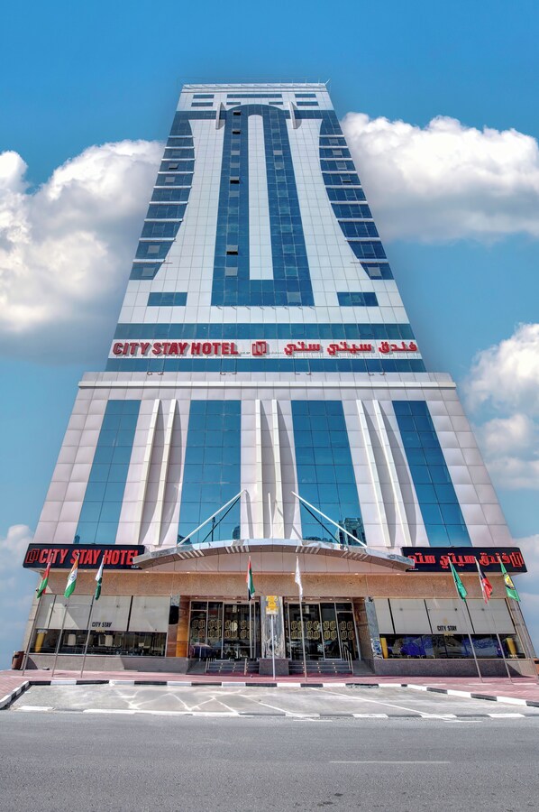 Un hotel alto con un diseño moderno, que destaca un cartel que dice "CITY STAY HOTEL" y varias banderas en la entrada.