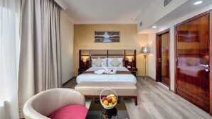 Una habitación de hotel con una cama grande, una zona de estar con una silla, una mesita de noche con lámpara y una cesta de frutas en el suelo.