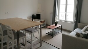 A modern living room with a wooden dining table, white chairs, a small black coffee table, a grey sofa, and a TV on a stand. - Nice apartment in the center of Touraine, in the heart of the Loire castles (L'Île-Bouchard)