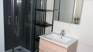 A modern bathroom with a glass-enclosed shower, a white sink with a wooden cabinet, and a black shelving unit. - Nice apartment in the center of Touraine, in the heart of the Loire castles (L'Île-Bouchard)