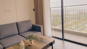 A modern living room with a grey sofa, a wooden coffee table with a plant, and a view of a cityscape through large windows. - 1BR 12FL-Vinhomes Ocean Park Ha Noi (Hanoi)