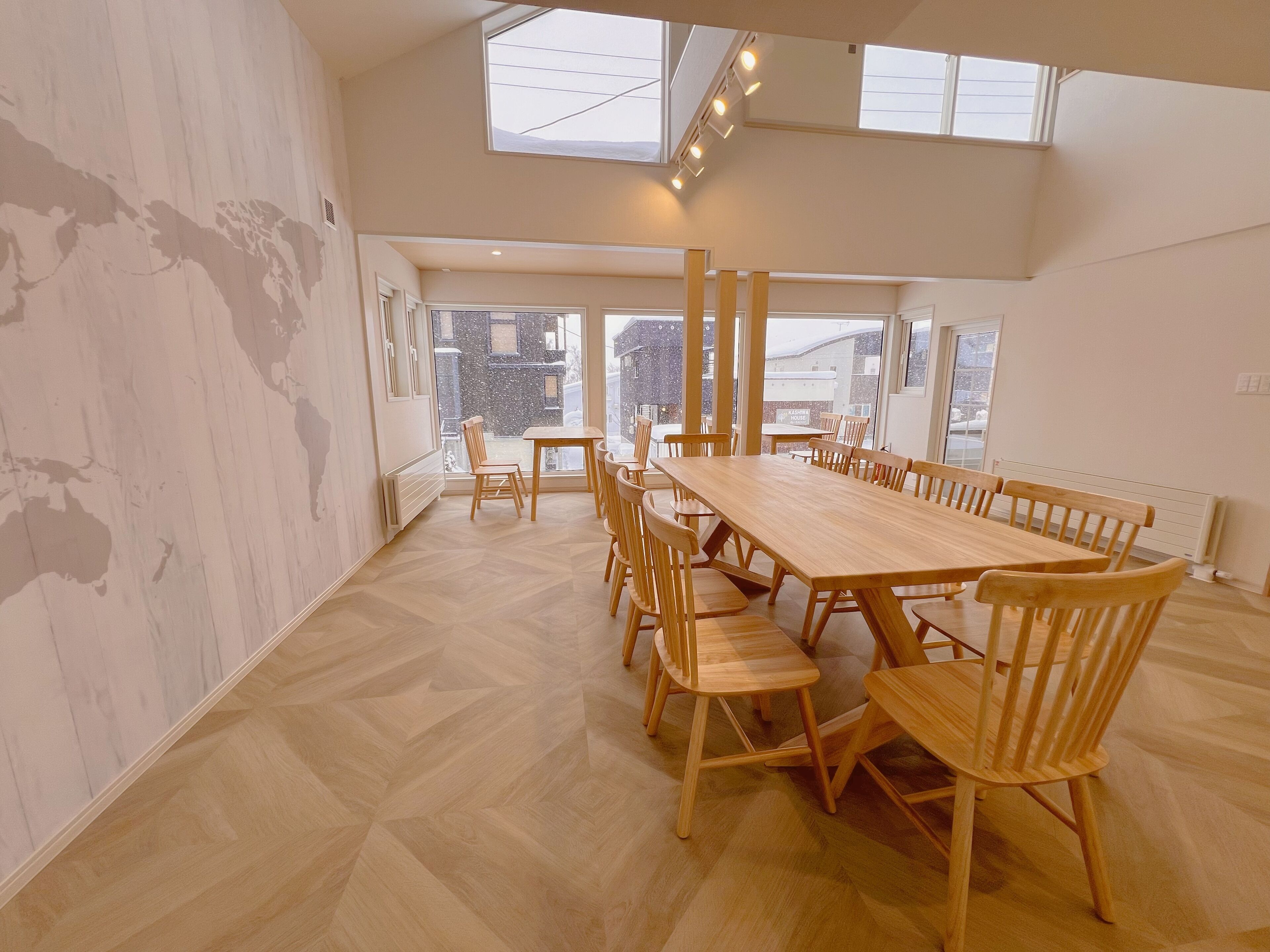 A spacious dining area with a long wooden table and chairs, large windows, and a high ceiling.