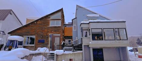 A two-story house with a wooden exterior and a modern extension with large windows. Snow covers the ground and steps.