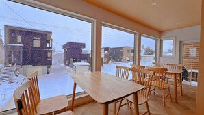 A dining area with a large table and chairs, large windows, and a view of a snowy street. - Snowball Hostel (Furano)